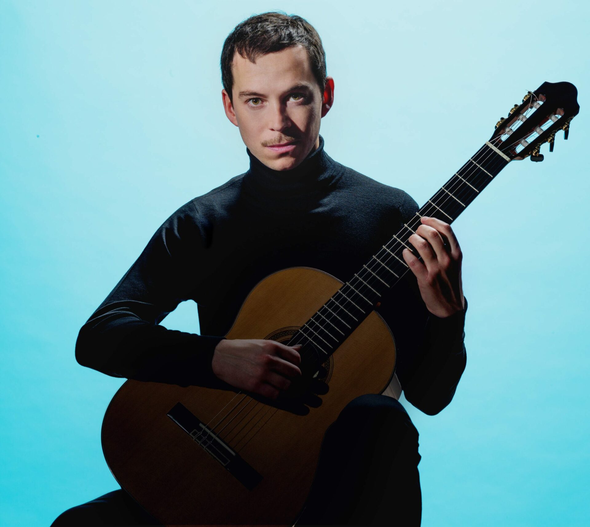 Thibault Cauvin est assis face à nous une guitare à la main et derrière lui se trouve un fond bleu ciel